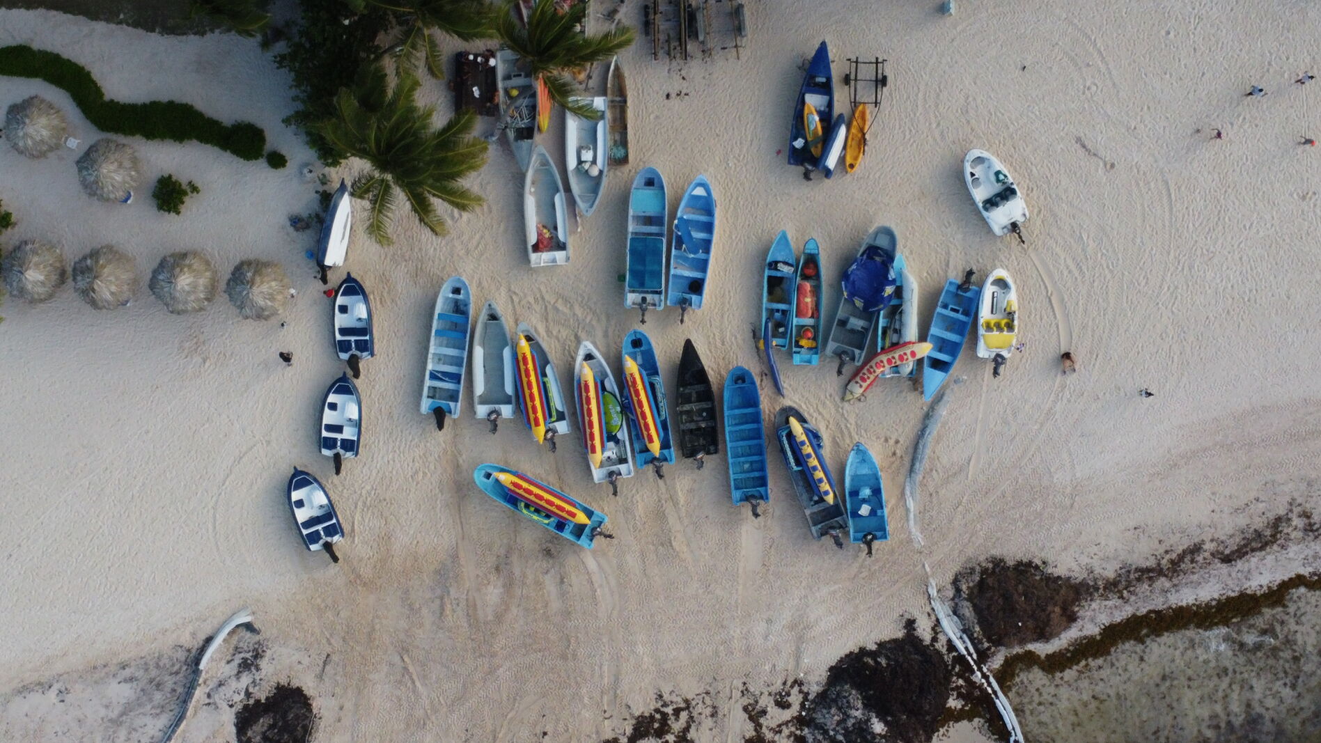 Top-down aerial of colorful pangas pulled onto a pale sand beach.